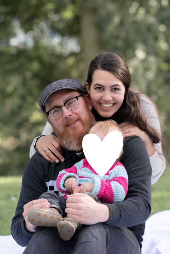 Foto einer dreiköpfigen Familie. Vorne das Kleinkind, dahinter sitzt Papa und dahinter steht kniend die Mama. Lächeln in die Kamera