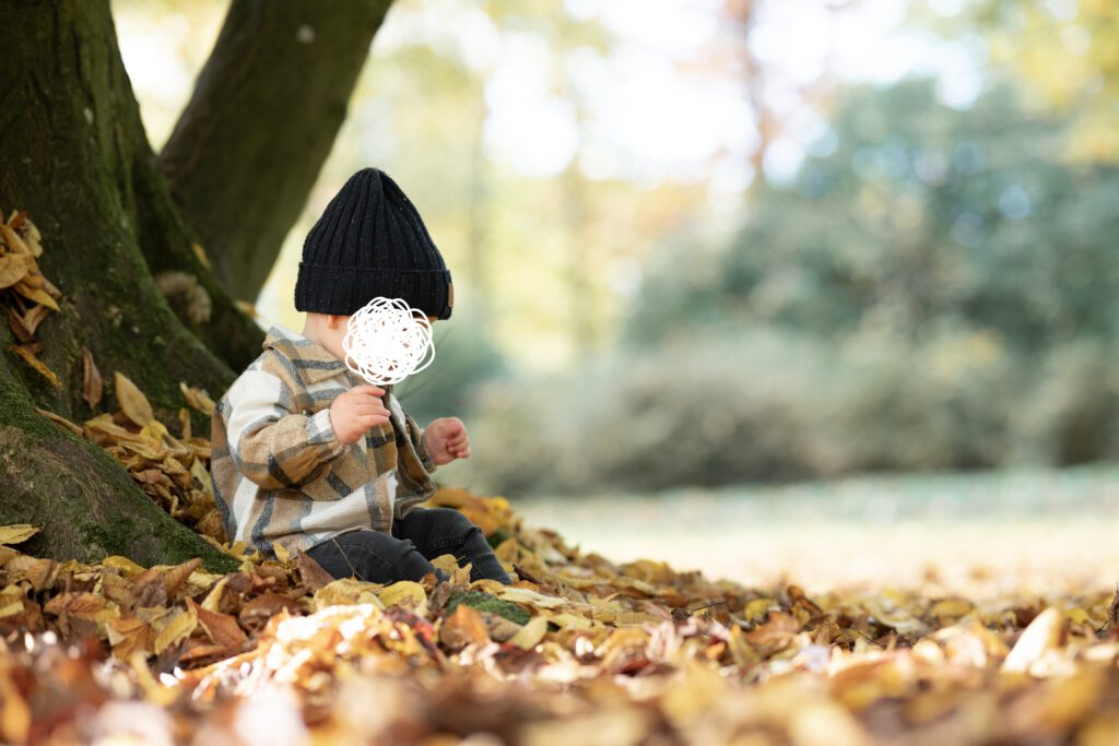 Bild eines Kleinkindes im Herbstlaub. Das Kind sitzt auf dem Laub an einem Baum gelehnt und trägt eine Mütze