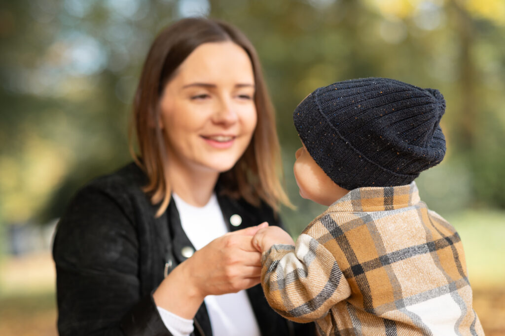 Foto von Mama und Kind. Die Mama hält das Kind an den Händen. Das Kind ist dem Fotografen mit dem Rücken zugewandt
