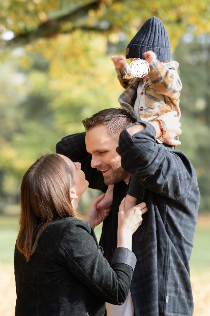 Foto einer dreiköpfigen Familie. Der Vater hat das Kleinkind auf den Schultern und die Mutter steht dem Vater gegenüber.