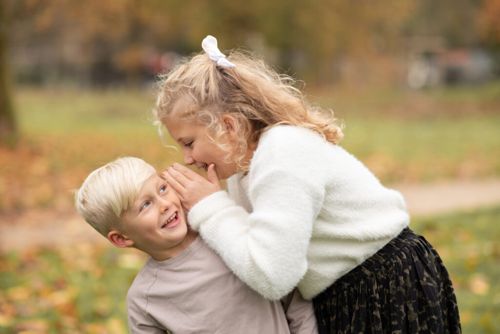 Foto zweier Geschwister. Das Mädchen flüstert dem Jungen etwas ins Ohr