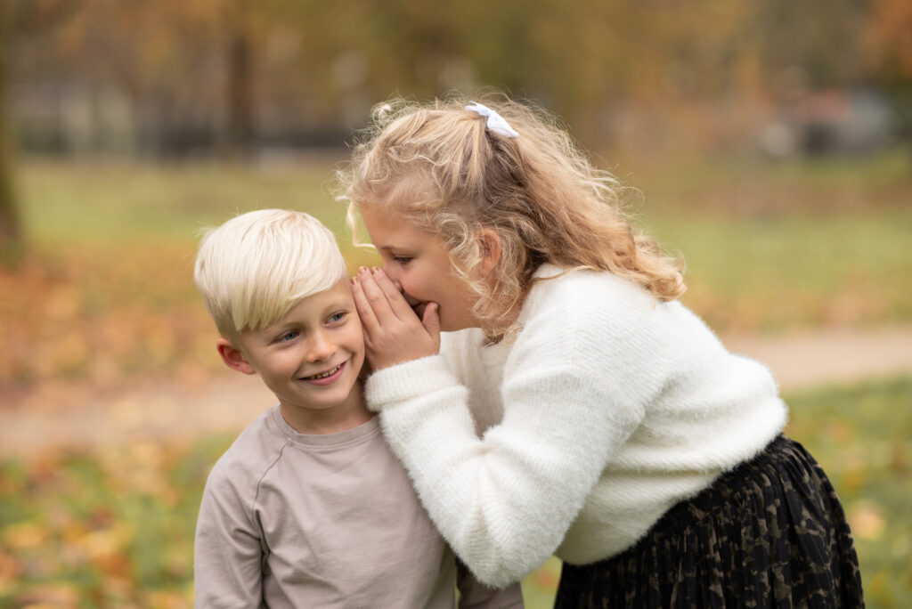 Bild zweier Geschwister. Das ältere Mädchen flüstert dem kleinen Jungen etwas ins Ohr. Buche dein Fotoshooting in Mönchengladbach