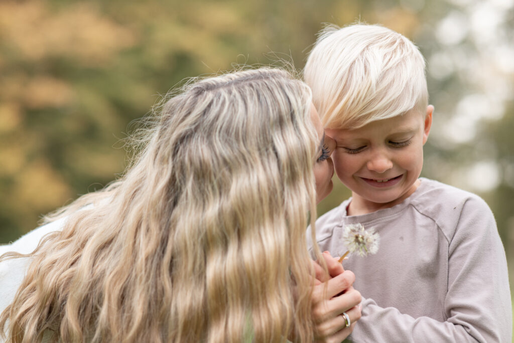 Mama küsst ihren Sohn auf die Wange. Er hält eine Pusteblume fest