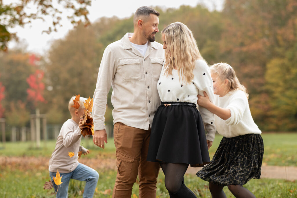 Bild einer vierköpfigen Familie im Herbstlaub. Die Kinder laufen um die Eltern herum, die in der Mitte stehen. Buche dein Fotoshooting in Mönchengladbach