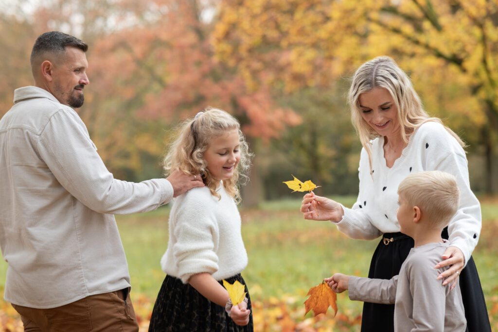 Foto einer Familie. Die zwei Kinder, Mädchen und Junge, sowie die Mama schauen, wer das schönste Herbstblatt in der Hand hält. Vater streicht der Tochter über die Schulter