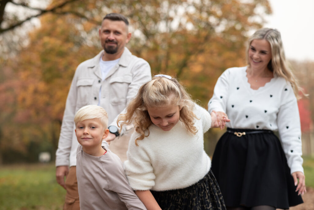 Foto einer vierköpfigen Familie. Alle halten sich an den Händen. Die Kinder, mittig, ziehen die Erwachsenen hinter sich her